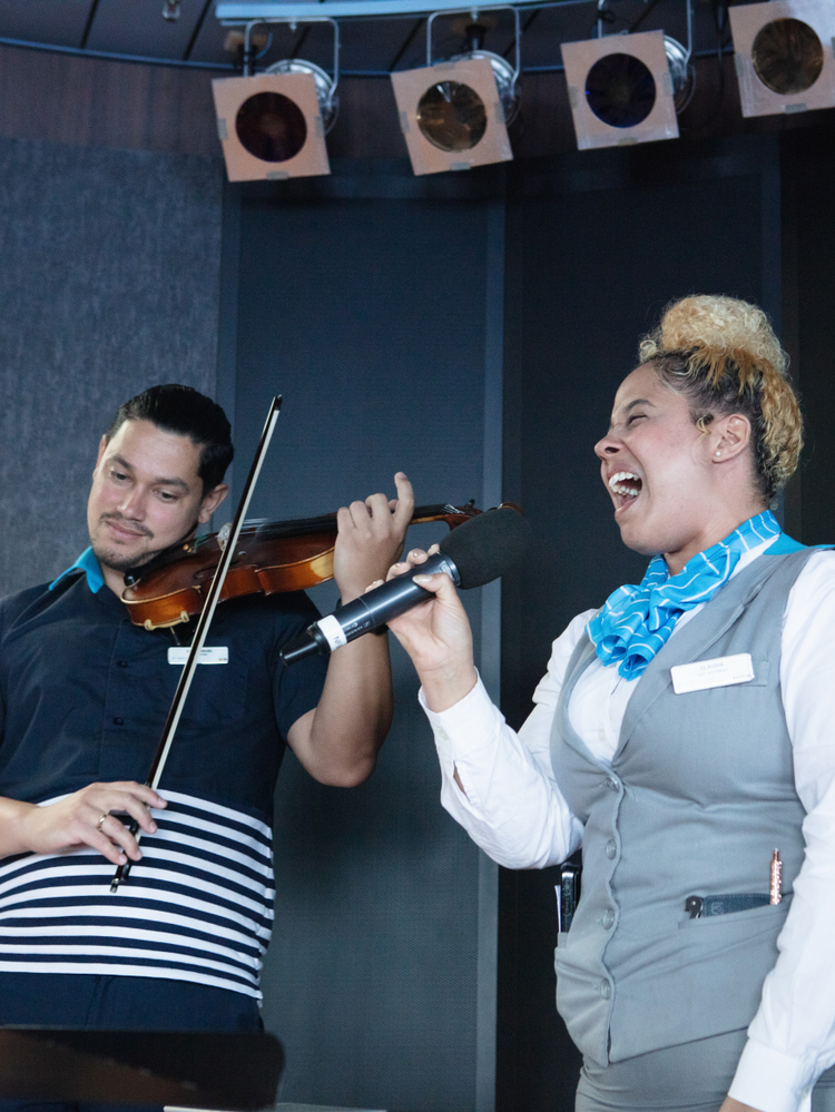 A member of Celestyal Journey crew sings into a microphone while another plays a violin
