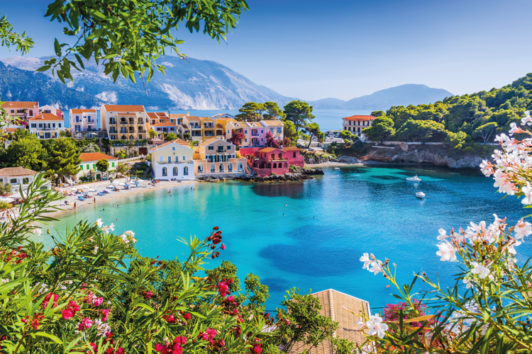 A photograph framed by flowers and plants lookin down into Assos Bay in Kefaloni. There are Mediterranean style houses in the mid-left-ground and views of the sea and mountains in the background