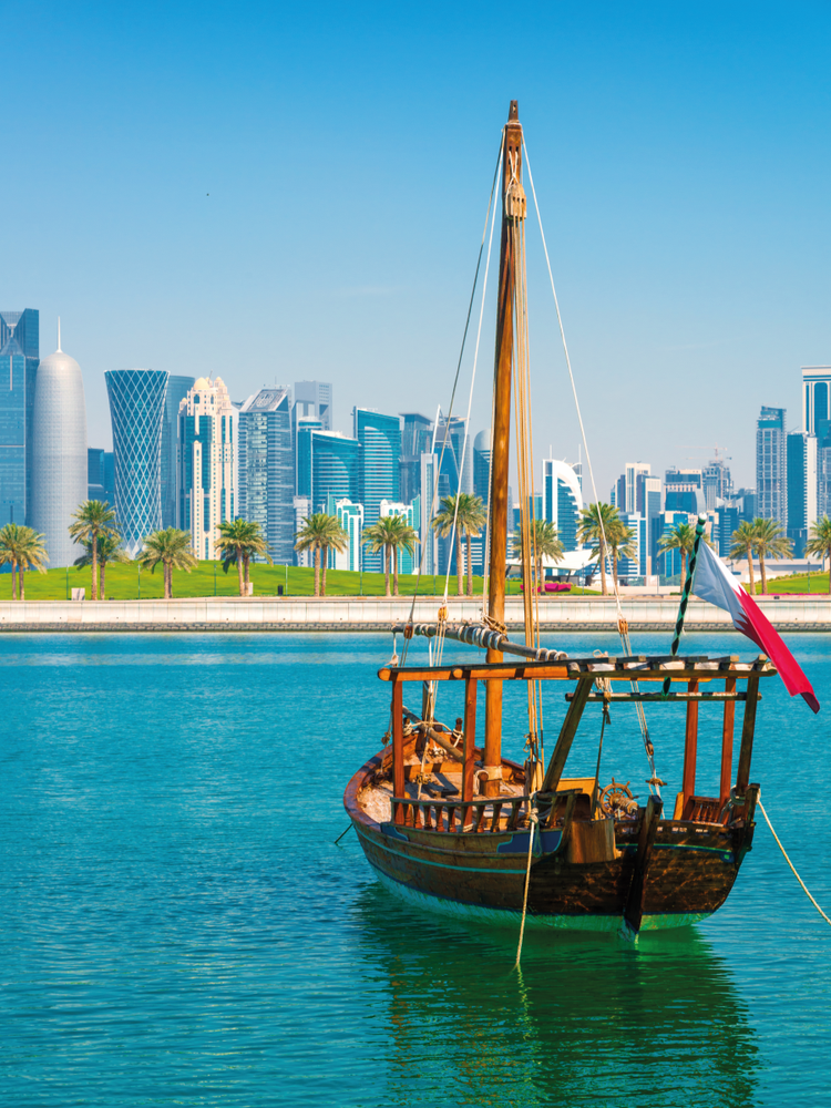 A photograph of an Arabian-style dhow boat in the sea with Doha&#39;s cityscape in the background