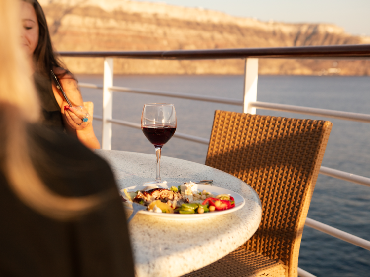 On the terrace of a cruise ship, a glass of red wine and plate of food on a table in the sun