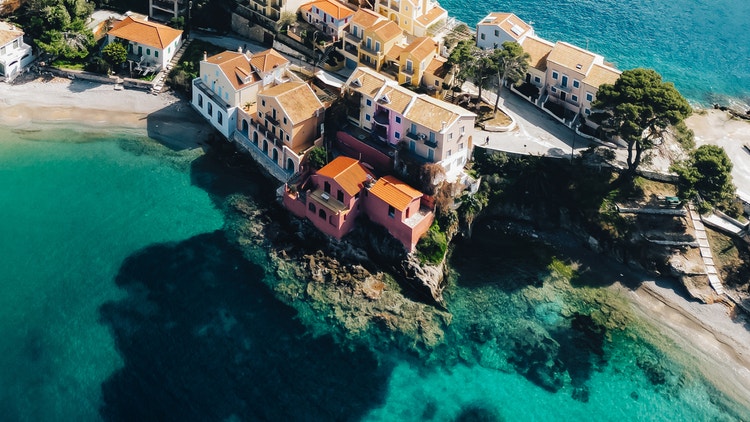 Vista aérea de coloridas casas de un pueblo mediterráneo con tejados de terracota, situadas dramáticamente sobre un promontorio costero rocoso, rodeadas de aguas turquesa cristalinas y vibrantes — descubre los icónicos destinos de las islas griegas con Celestyal Cruises.