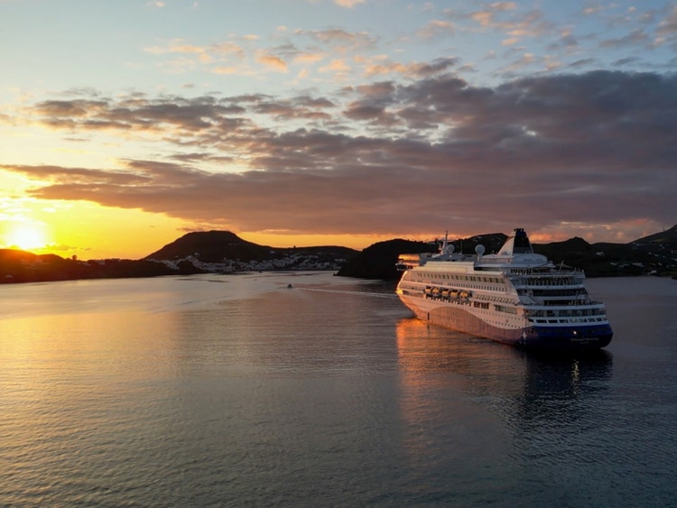 The Celestyal Discovery ship anchored in the bay of the Greek island port of Patmos at sunset, surrounded by the silohuette of the island&#39;s hilly terrain.