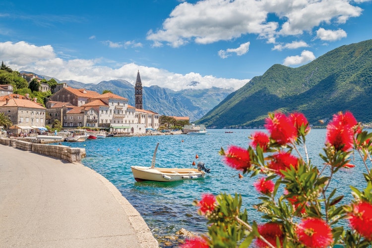 Image of a waterside path a bay with a small boat in the water, and a Montengran town in the background and mountains on either side.