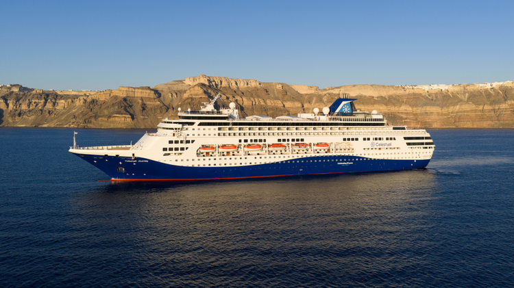 External shot of the Celestyal Journey at sea, with a mountainous coastline in the background