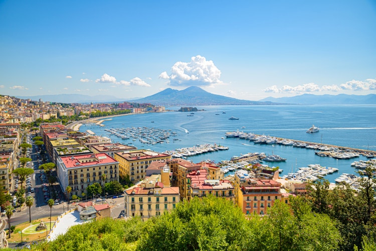 An elevated panoramic view of the Gulf of Naples, Italy, featuring a bustling harbor filled with boats and yachts, coastal city buildings, and Mount Vesuvius rising in the background under a clear blue sky.