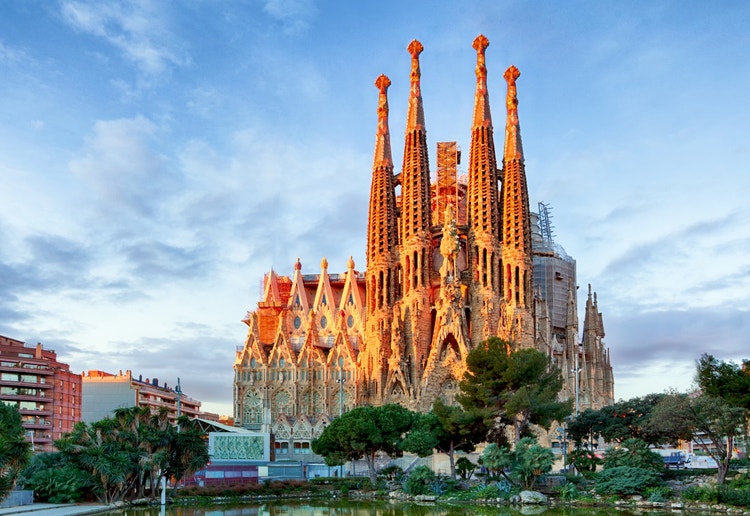 The Sagrada Família in Barcelona, Spain, seen from across the pond at Plaça de Gaudí, showcasing its towering spires and the Nativity Façade under a soft blue sky.
